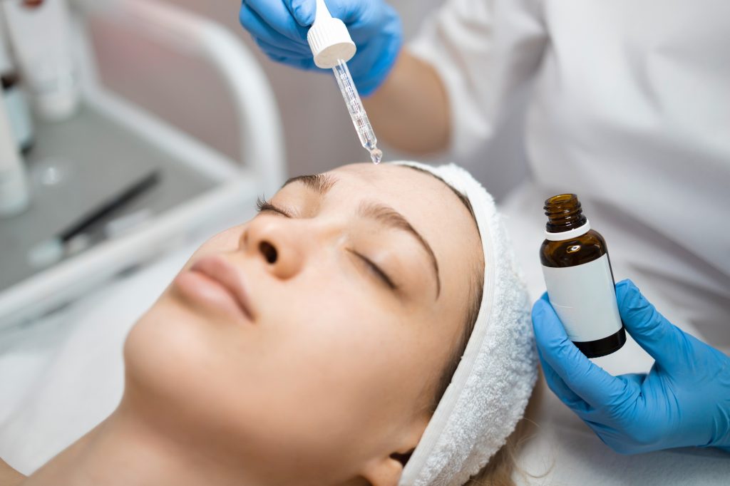 Hands of beautician dripping nourishing oil on forehead and facial skin of young woman